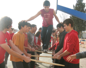 לימודי חינוך גופני בתמונה ילדים עושים גשר ממקלות בזמן שילד אחד מטפס על המקלות