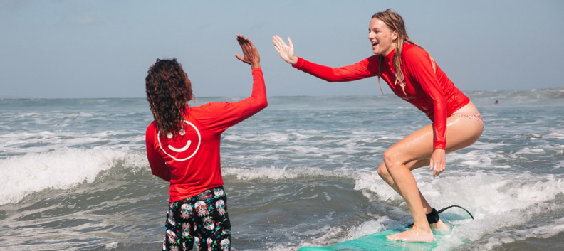 קורס מדריכי גלישת גלים בתמונה אישה על גלשן תופסת גל ונותנת ומנופפת לשלום למדריך גלישה